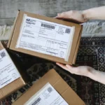 Tangan memegang paket kardus dengan label pengiriman yang menampilkan nomor waybill